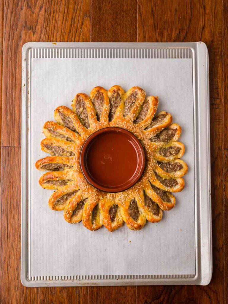 Golden baked sausage roll wreath on a baking sheet with the bowl still in the center for cranberry sauce.