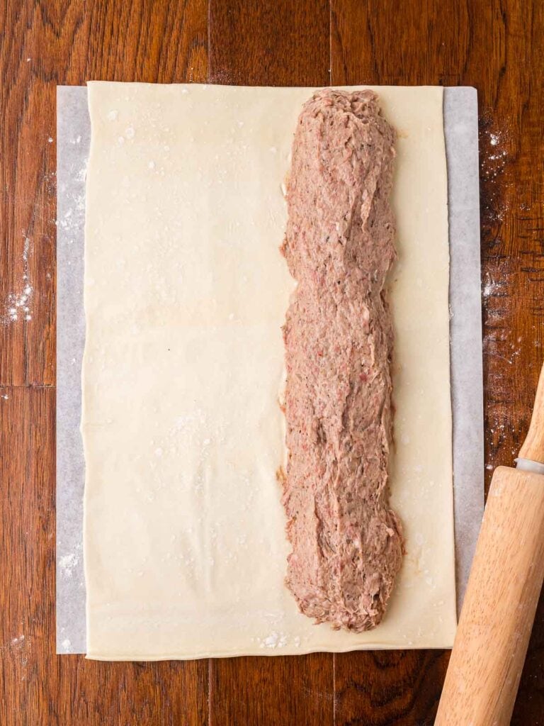 Puff pastry sheet with a long line of sausage filling spread along one edge.