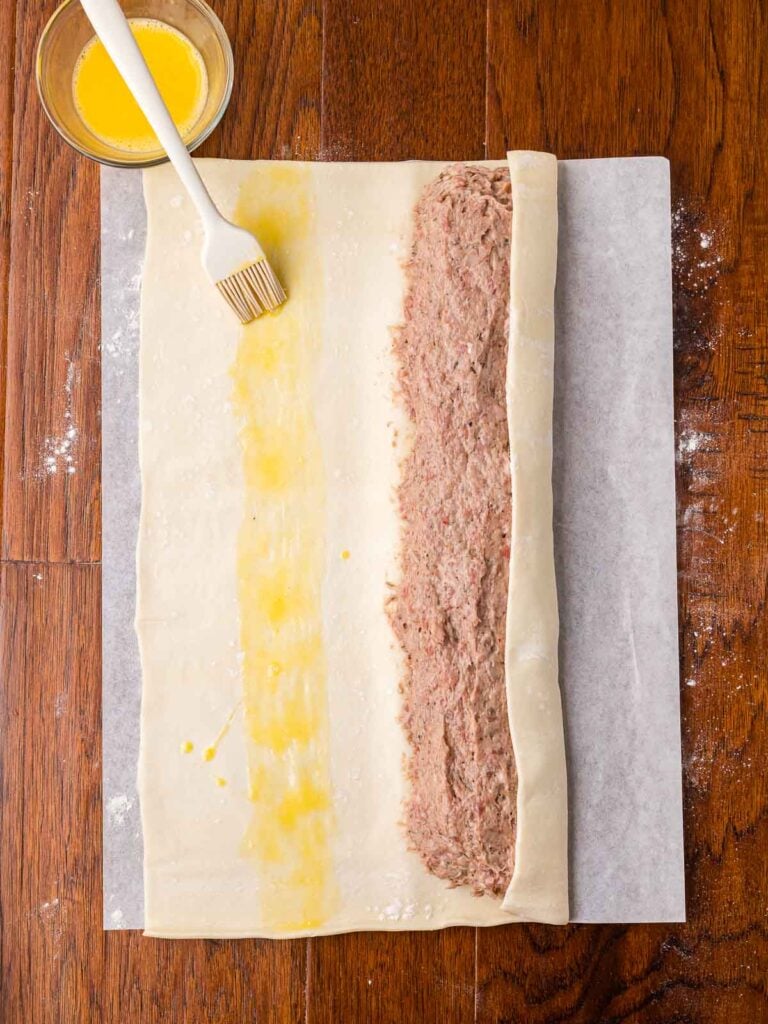 Brushing egg wash on puff pastry before rolling to seal the sausage filling.