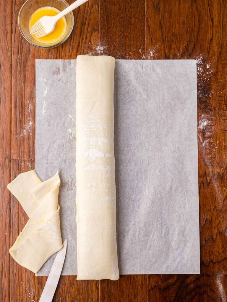 Rolled puff pastry sausage log with trimmed edges and egg wash ready to shape.