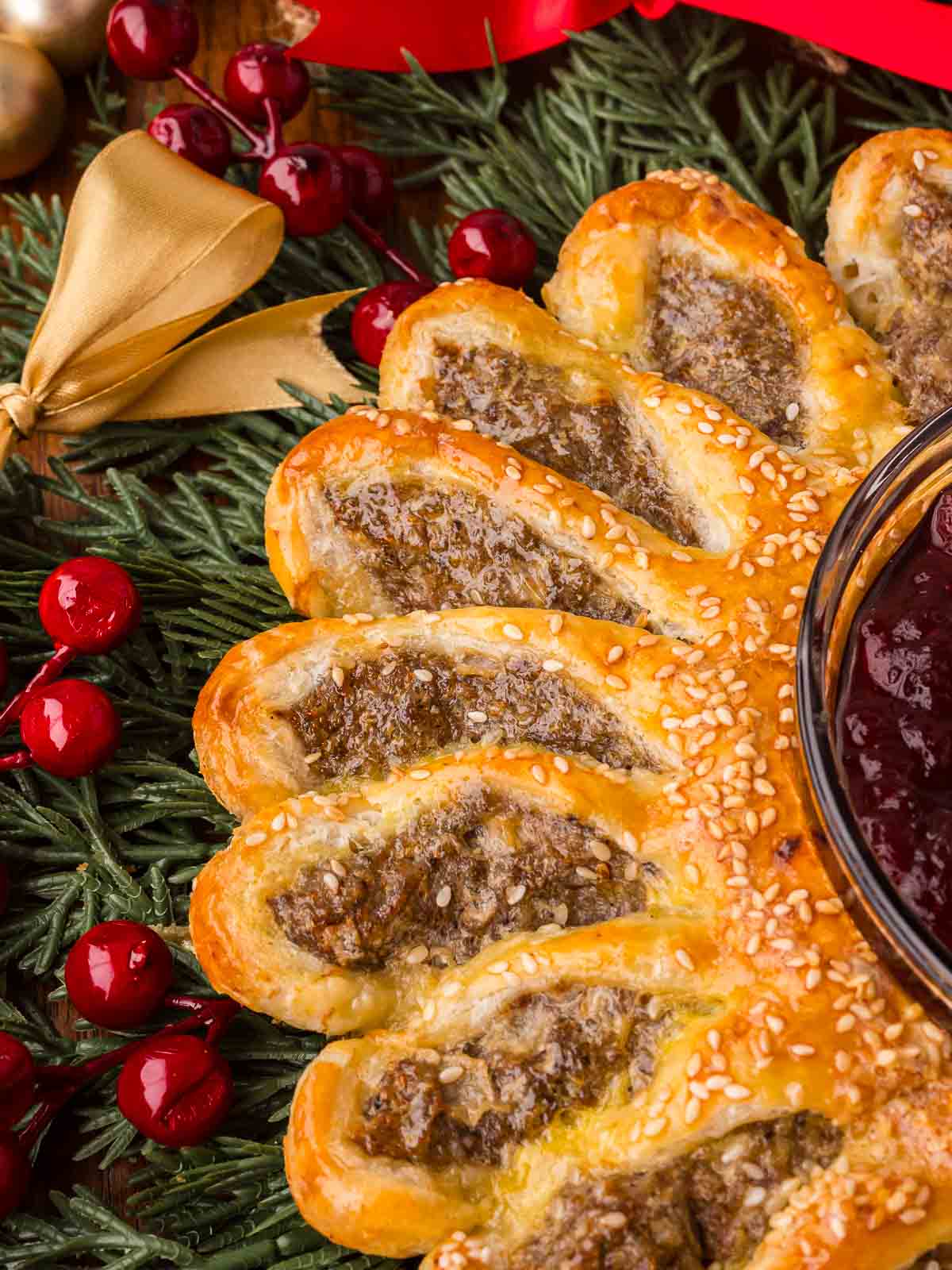Sausage roll wreath with flaky pastry and cranberry sauce in the center, decorated with greenery and red ornaments.