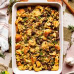 Full baking dish of sourdough stuffing with sausage and herbs, surrounded by fresh thyme and rosemary.