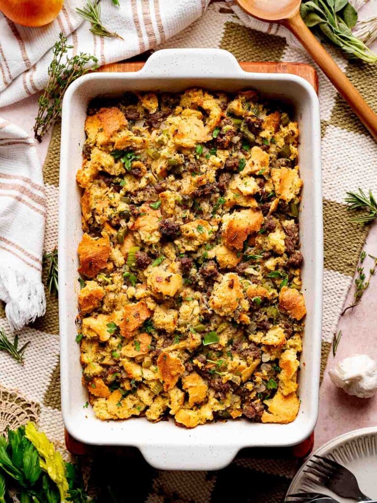 Full baking dish of sourdough stuffing with sausage and herbs, surrounded by fresh thyme and rosemary.