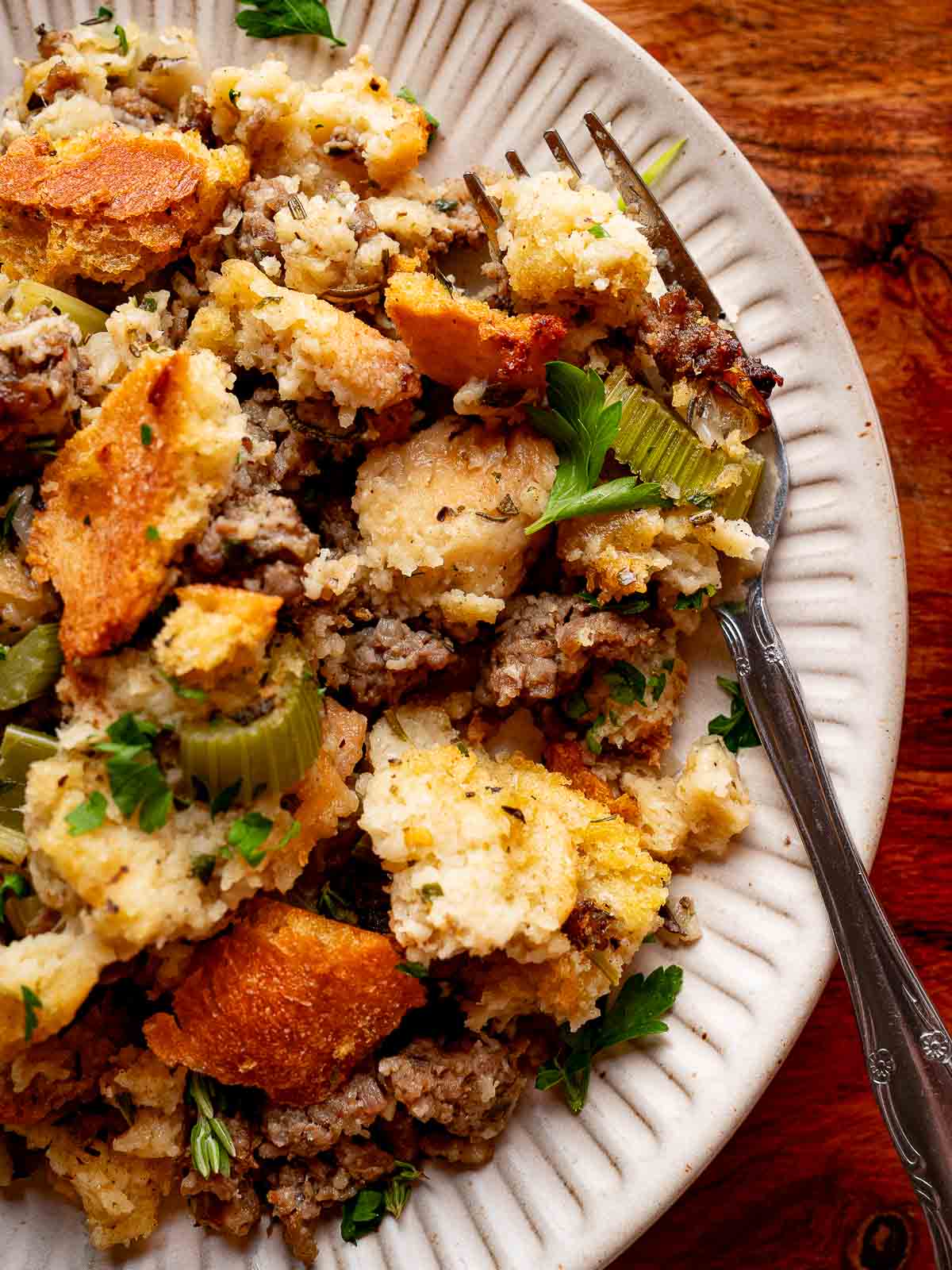Close-up of baked sourdough stuffing with sausage, celery, and herbs served on a white plate with a fork.