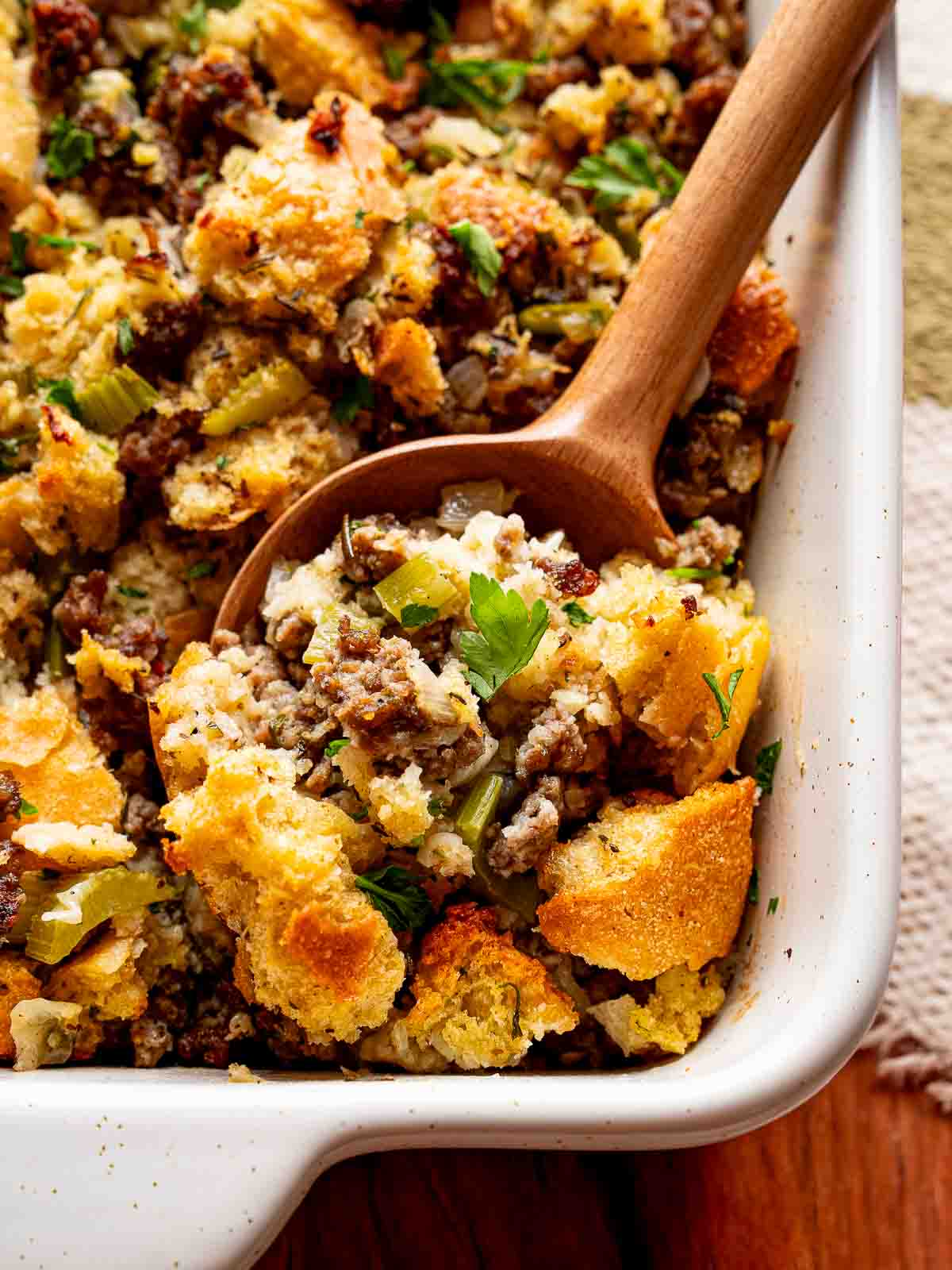 Wooden spoon scooping out sourdough stuffing with sausage, celery, and herbs from a baking dish.