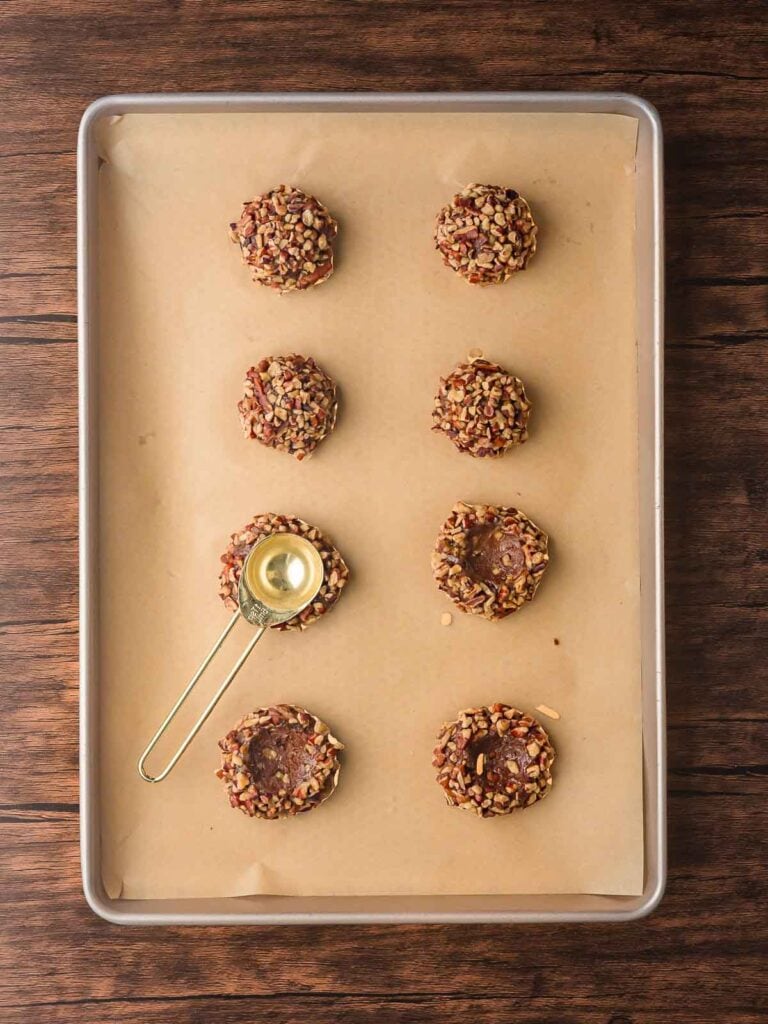 Unbaked pecan-coated cookie dough with thumbprints pressed in the center using a measuring spoon.