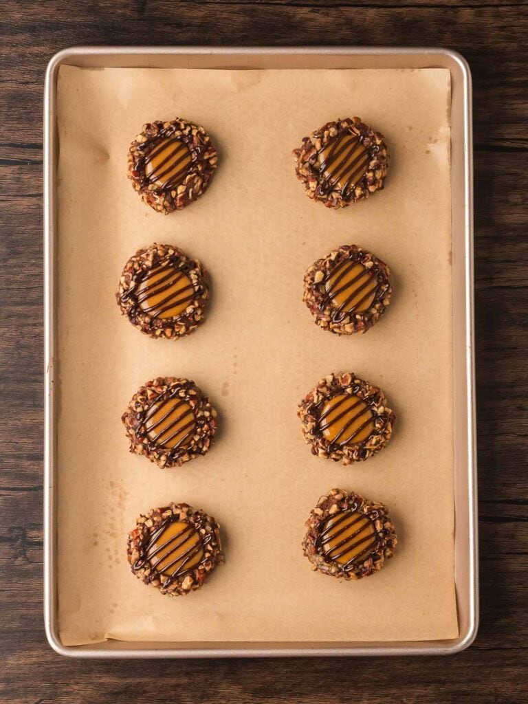 Turtle Thumbprint Cookies drizzled with melted chocolate on a parchment-lined baking sheet.