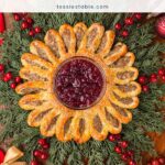 Overhead view of a golden sausage roll wreath with cranberry sauce in the center on evergreen branches with red berries by Tessie's Table.