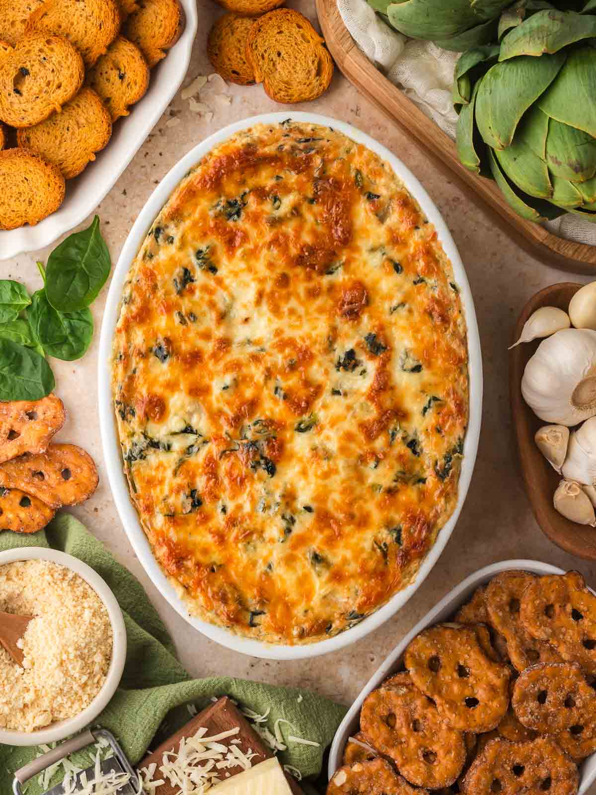 Baked spinach artichoke dip surrounded by baguette slices, pretzels, and ingredients.