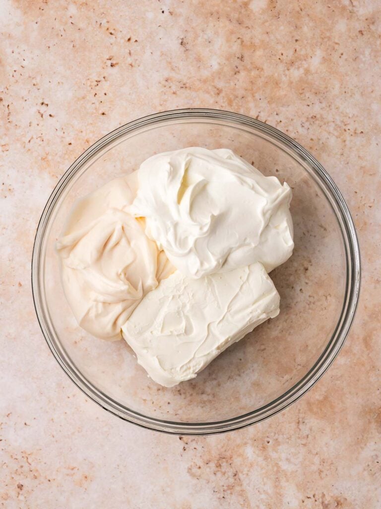 Cream cheese, sour cream, and mayonnaise in a glass bowl on a light countertop.