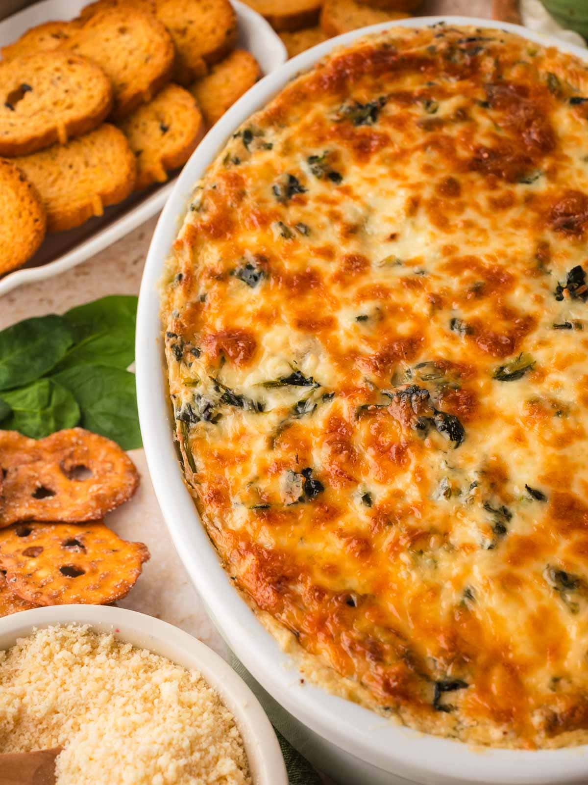 Spinach artichoke dip with pretzels, bread slices, parmesan, and fresh spinach around the dish.