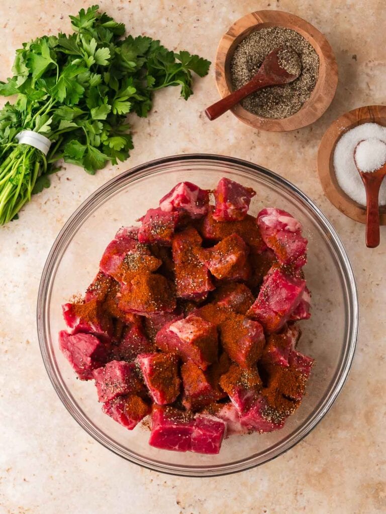 Bowl of raw steak pieces seasoned with smoked paprika, salt, and pepper, surrounded by parsley and small bowls of seasoning.