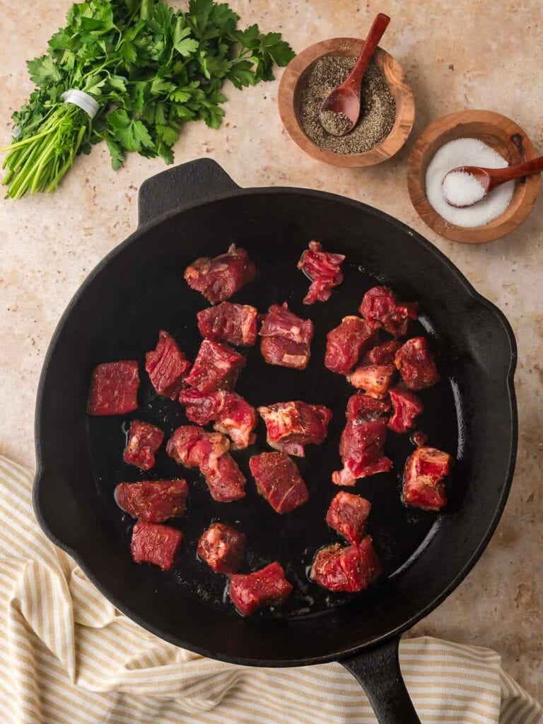 Cast iron skillet with the first batch of raw seasoned steak bites searing on the surface.