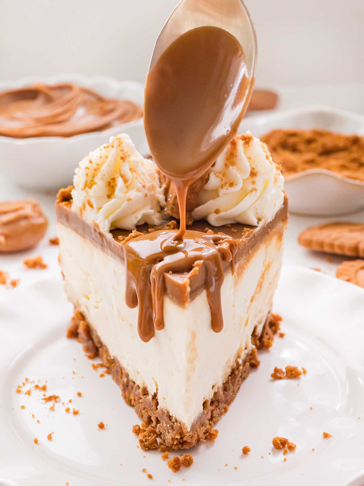 Spoon drizzling warm Biscoff cookie butter over a slice of cheesecake topped with whipped cream.