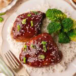 Overhead shot of honey garlic chicken on a white plate with broccoli and rice, surrounded by small bowls of green onions and dumplings.
