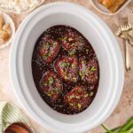 Slow cooker filled with glossy honey garlic chicken thighs garnished with sesame seeds and green onions, surrounded by bowls of rice, egg rolls, and dumplings.