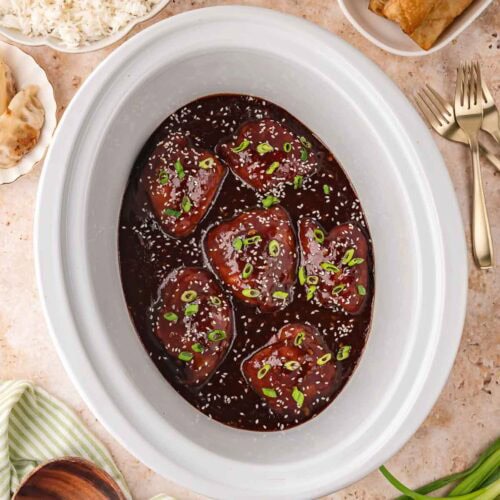 Slow cooker filled with glossy honey garlic chicken thighs garnished with sesame seeds and green onions, surrounded by bowls of rice, egg rolls, and dumplings.