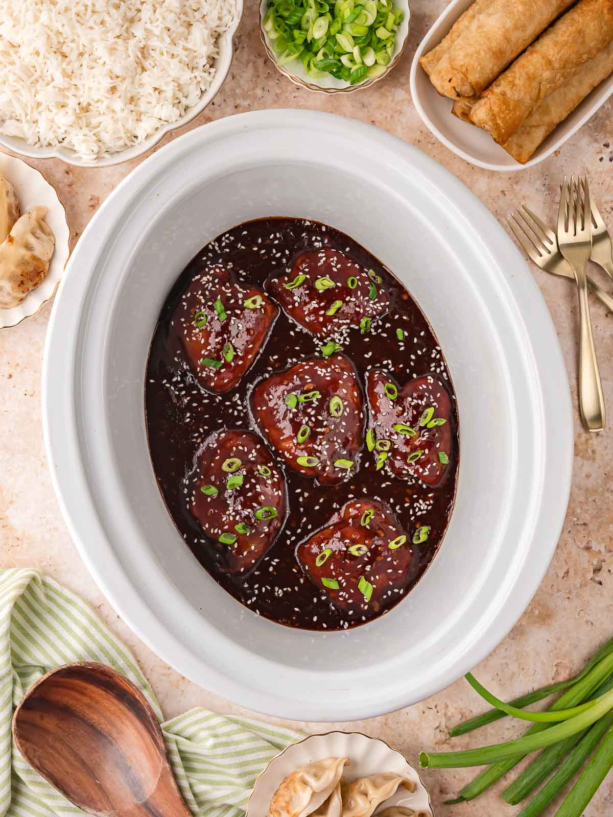 Slow cooker filled with glossy honey garlic chicken thighs garnished with sesame seeds and green onions, surrounded by bowls of rice, egg rolls, and dumplings.