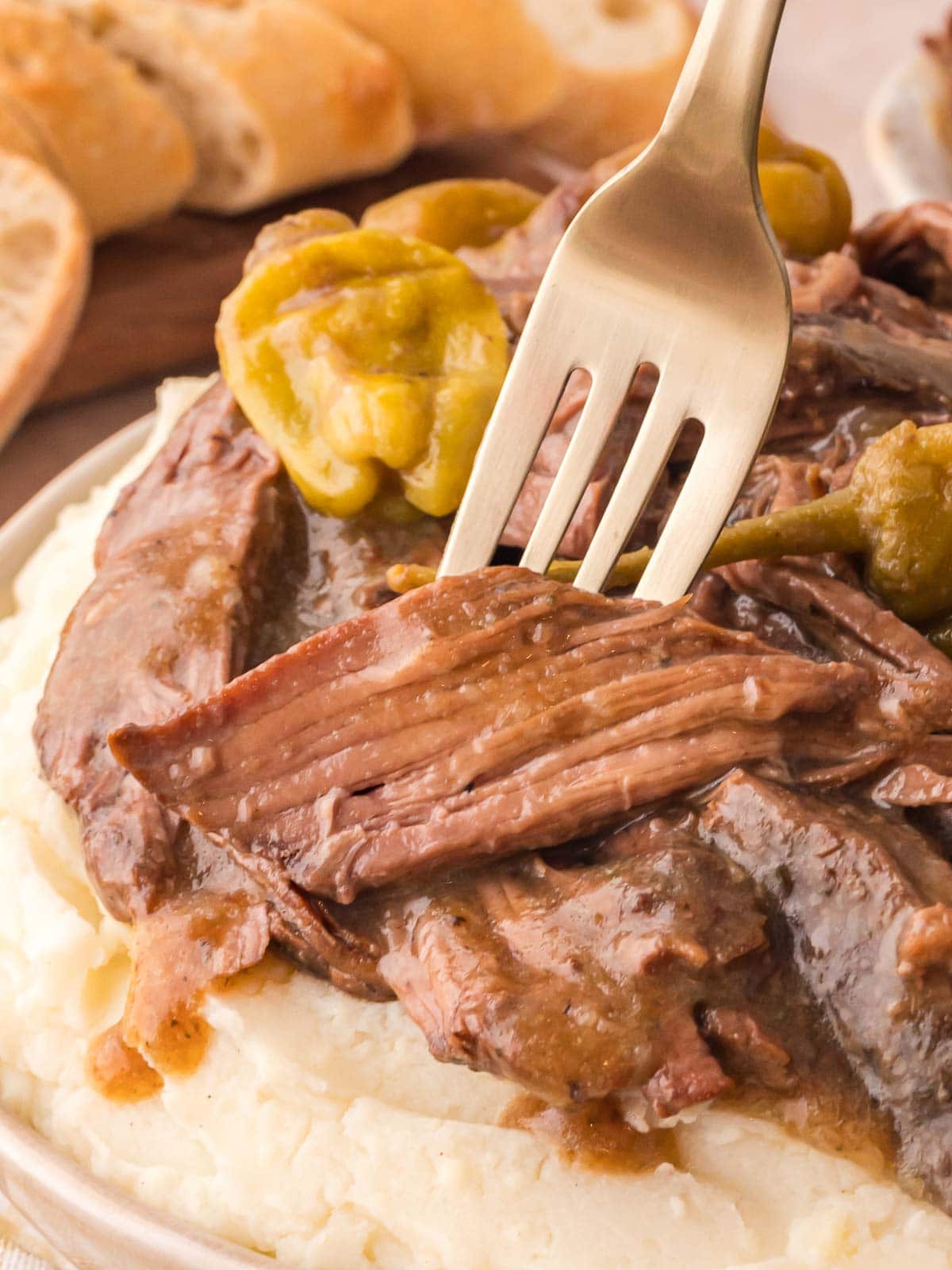 Fork pulling a piece of tender Mississippi Pot Roast from a plate of mashed potatoes with pepperoncini on top.