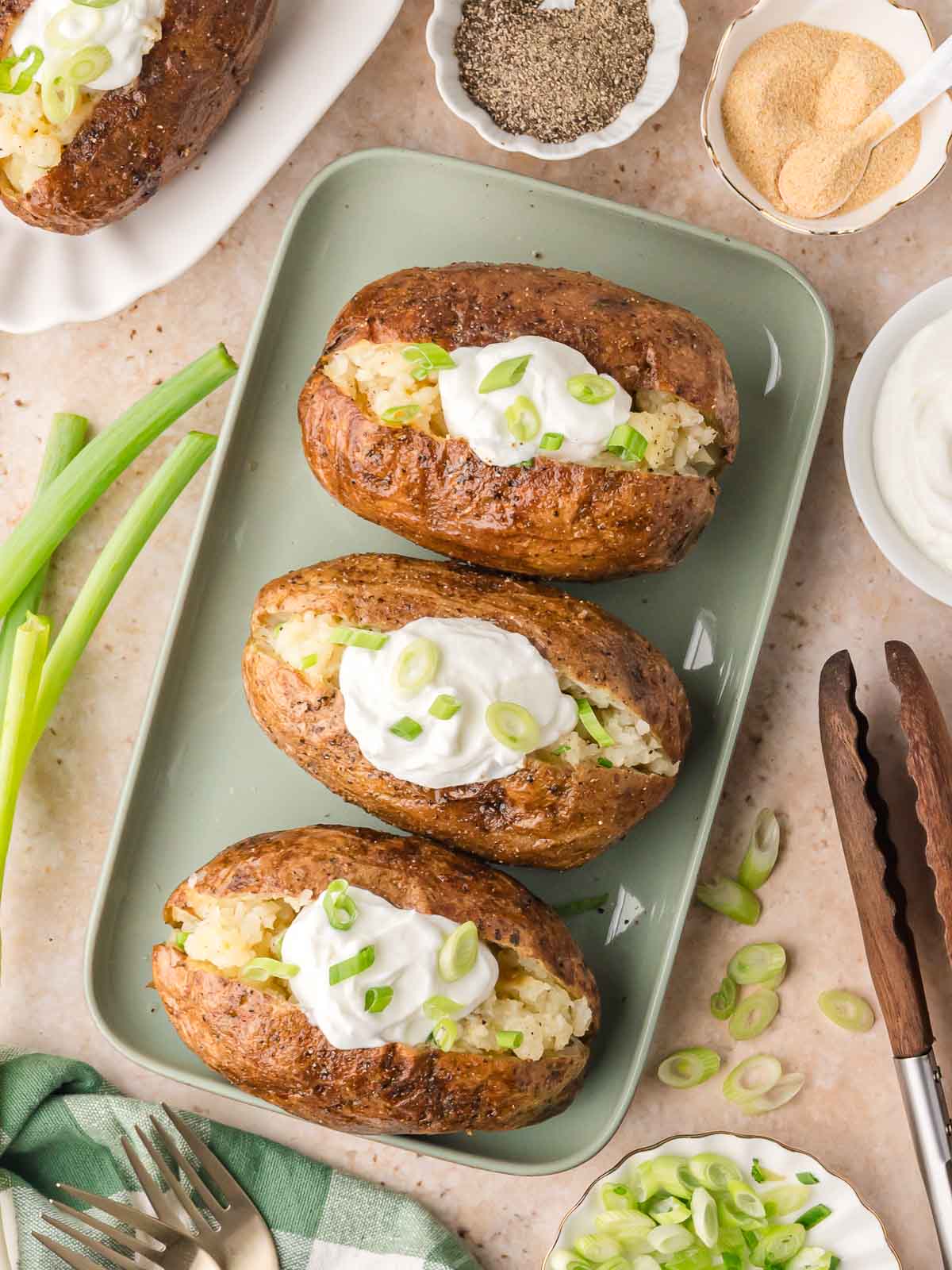 Overhead view of baked potatoes topped with sour cream and green onions, surrounded by small bowls of seasonings and green onions.