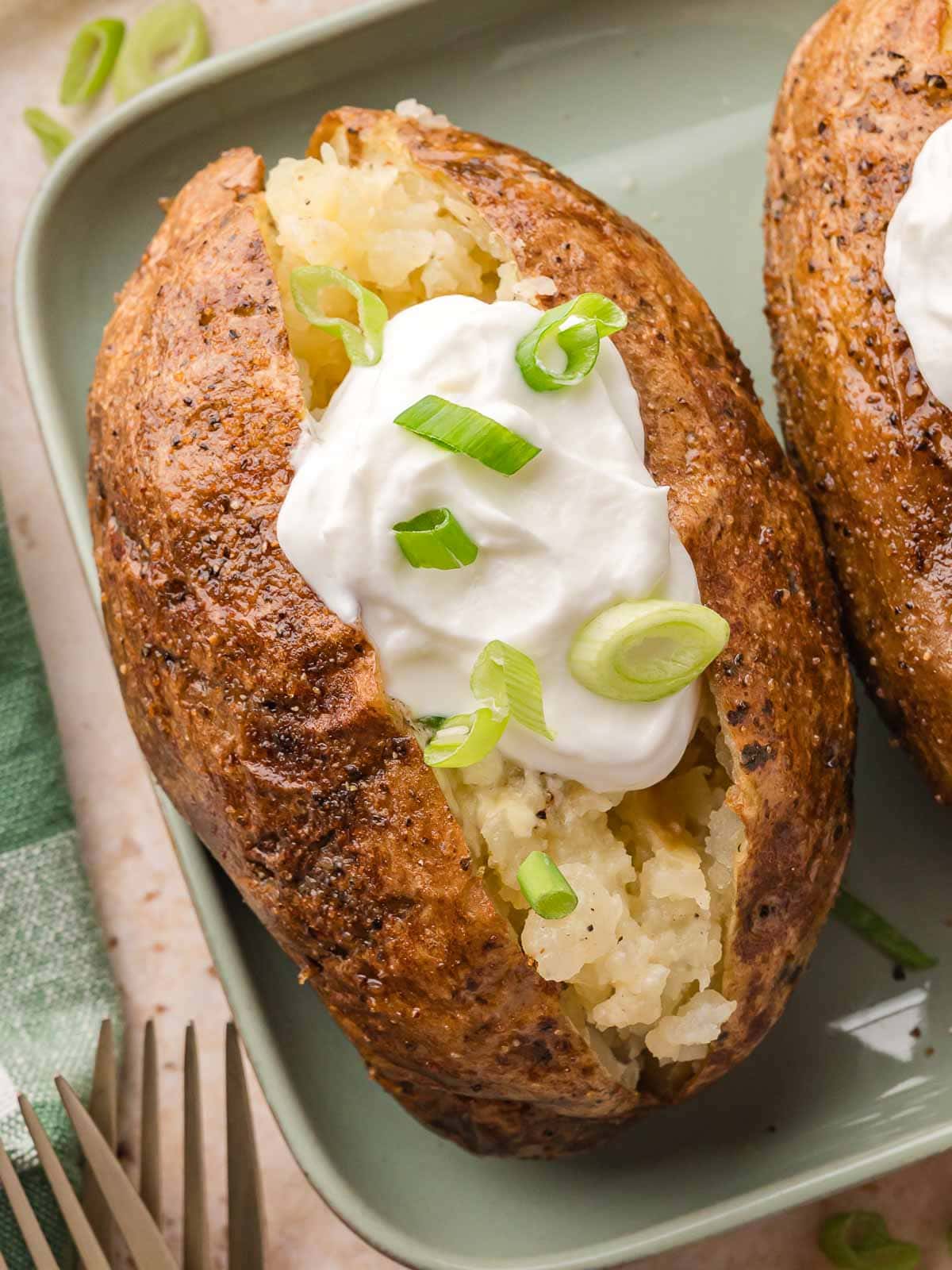 Baked potato filled with fluffy potato and topped with sour cream and green onions, served on a green plate next to a fork.