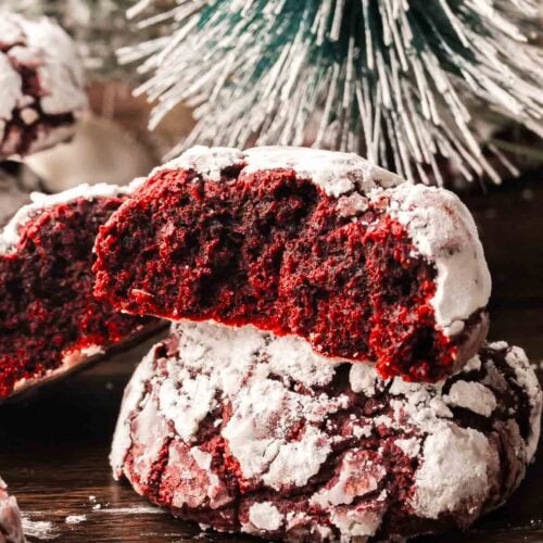 Red velvet crinkle cookie broken in half to show its soft, chewy inside with snowy powdered sugar coating, set in front of a frosted Christmas tree.