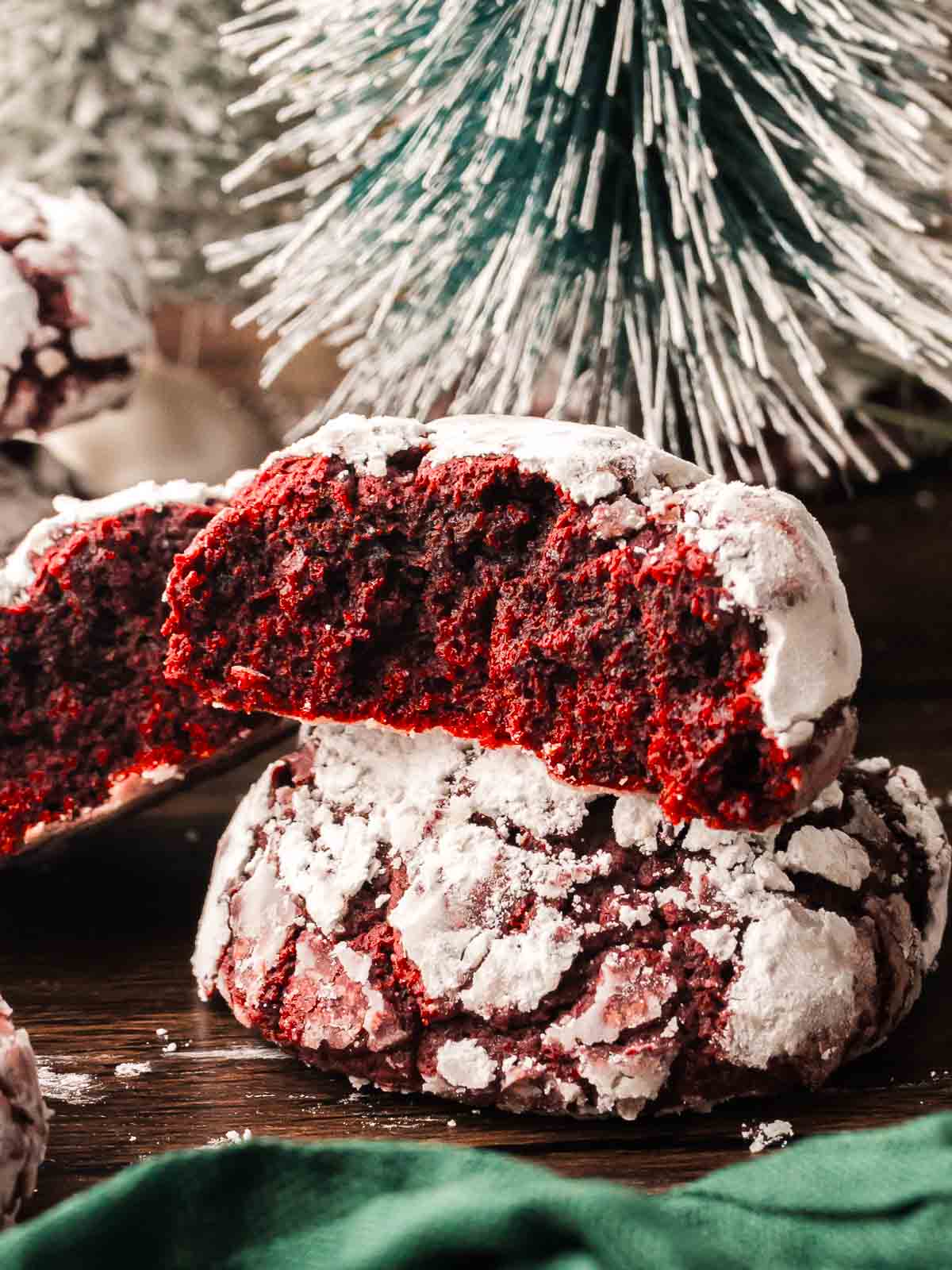 Red velvet crinkle cookie broken in half to show its soft, chewy inside with snowy powdered sugar coating, set in front of a frosted Christmas tree.