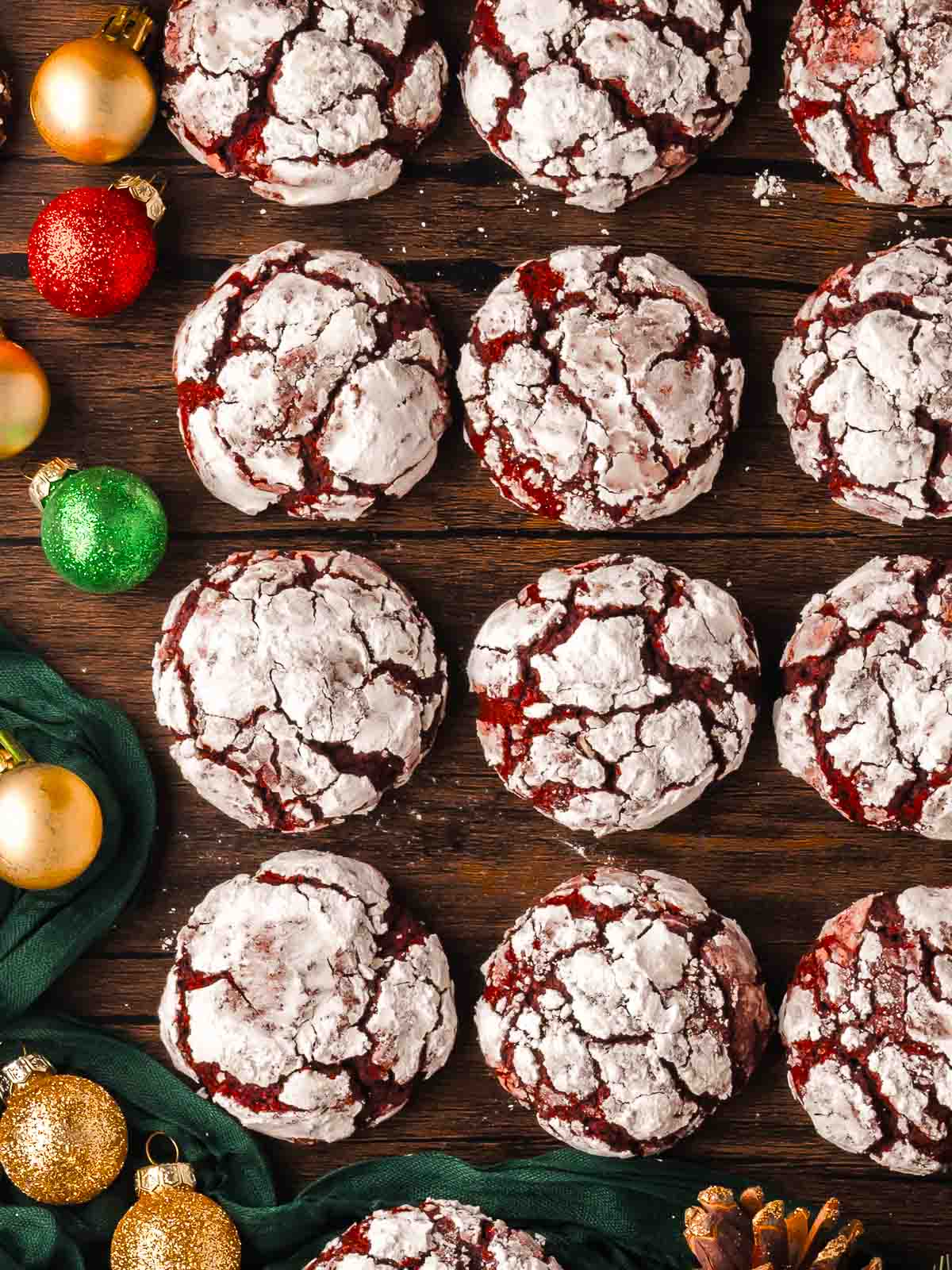 Rows of red velvet crinkle cookies coated in powdered sugar on a wooden surface with gold, red, and green ornaments for a festive Christmas look.