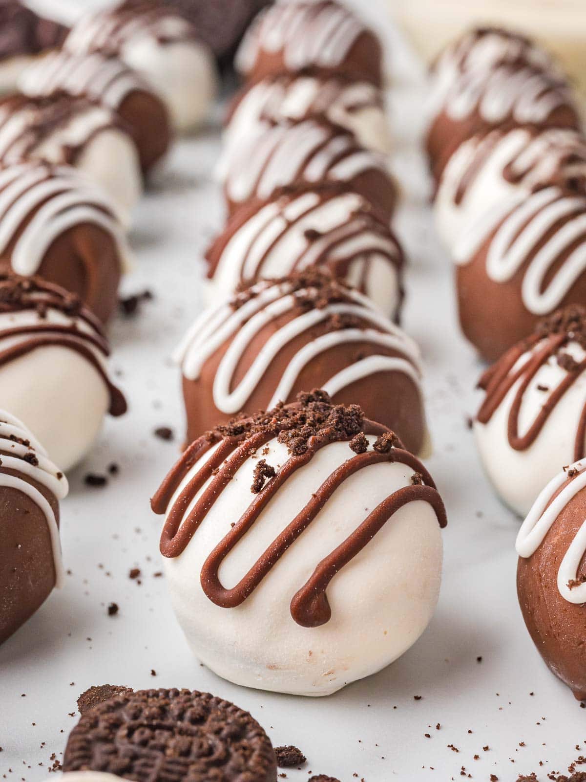 Rows of decorated Oreo truffles drizzled with chocolate and sprinkled with Oreo crumbs on a white surface.