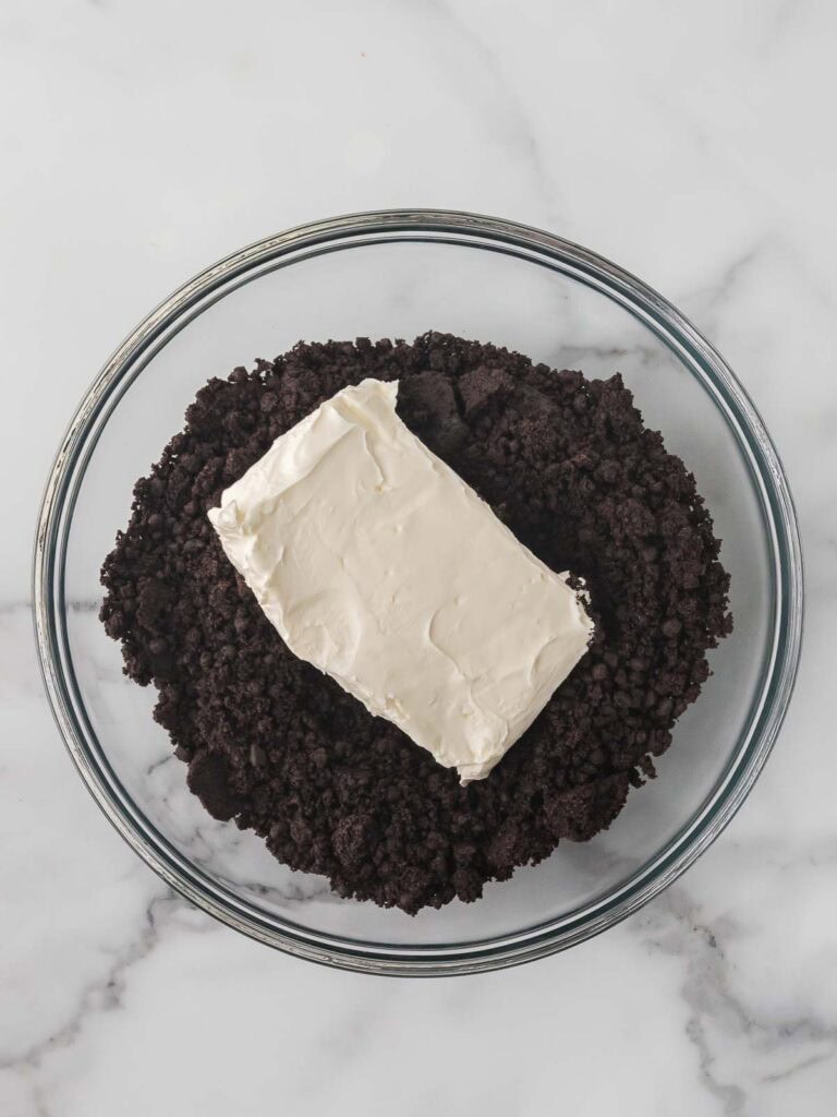 A block of softened cream cheese sitting on top of a bowl of crushed Oreo crumbs before mixing to form the truffle filling.