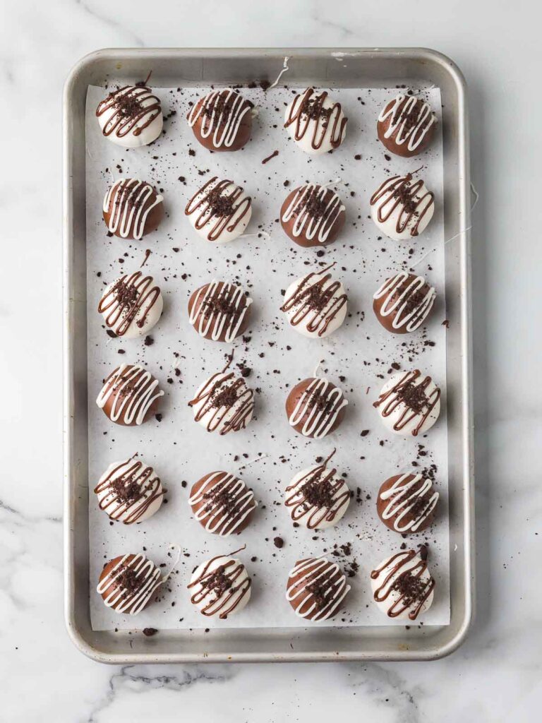 Finished Oreo truffles drizzled with chocolate and topped with crushed Oreo crumbs on a parchment lined baking sheet.