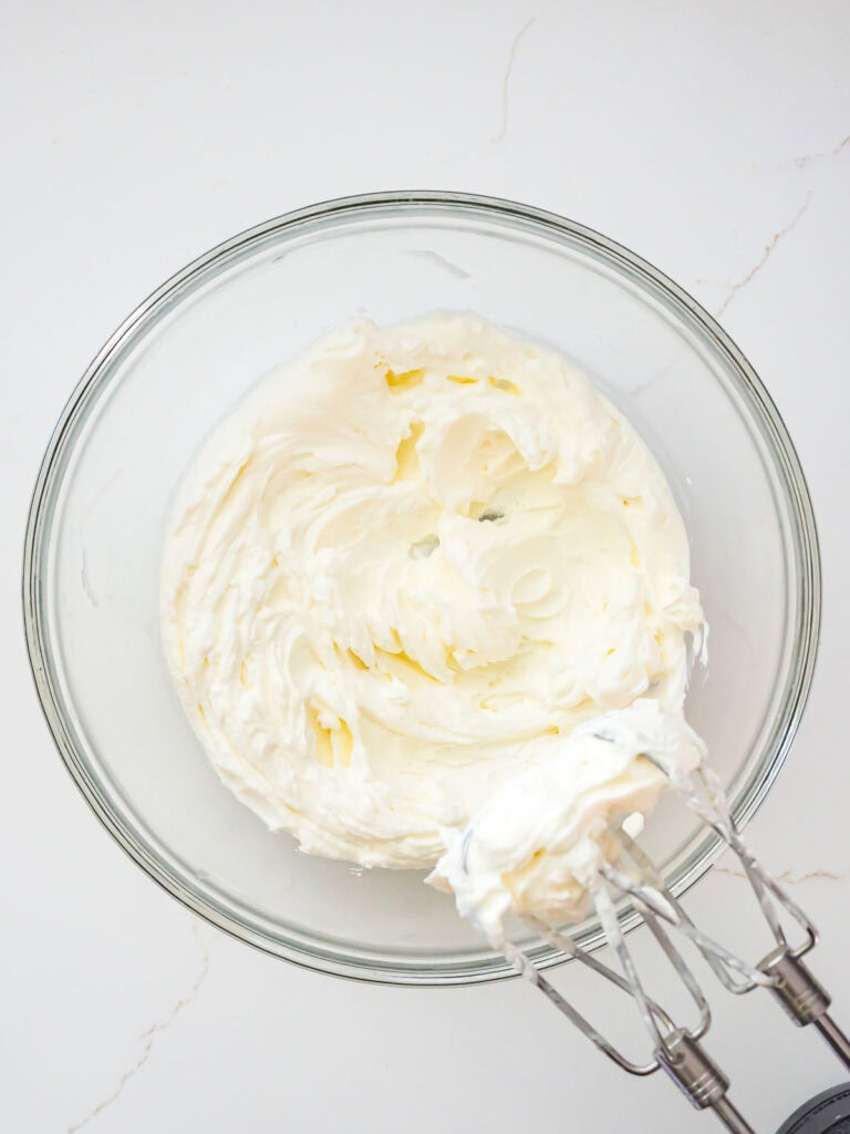 Cream cheese mixture being beaten with a hand mixer until creamy and lump-free.