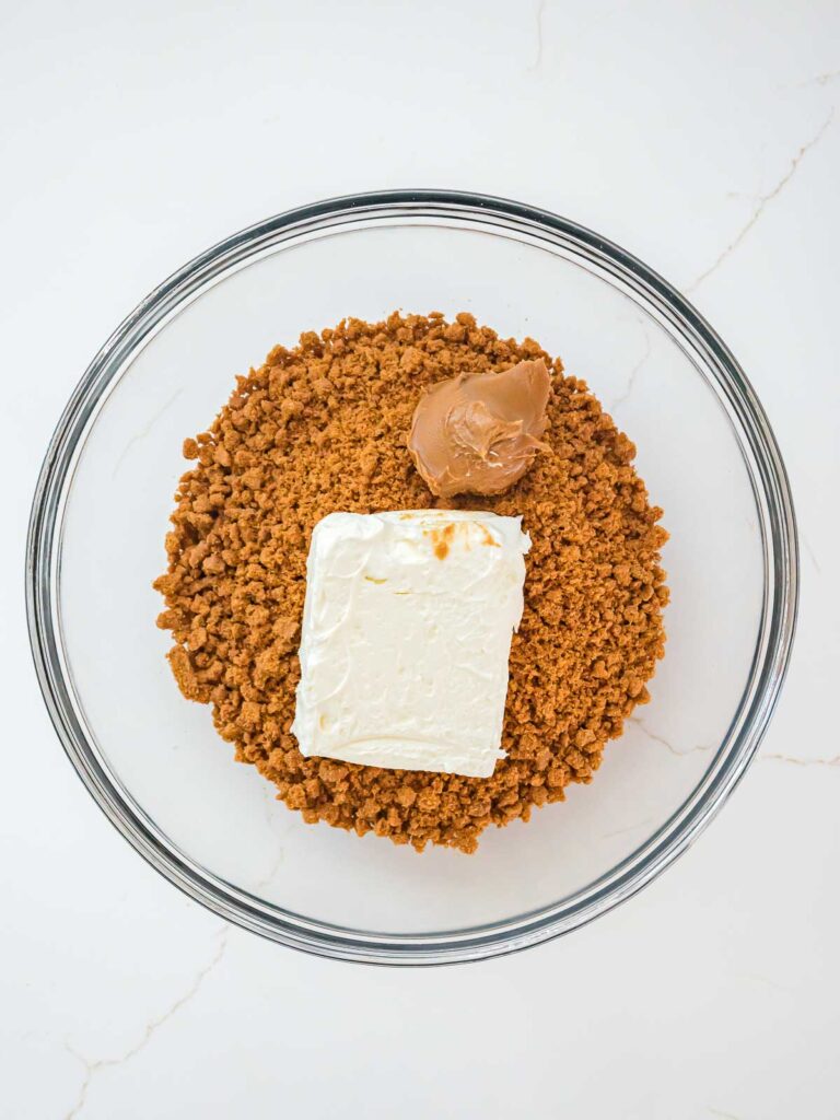 Crushed Biscoff cookies, softened cream cheese, and cookie butter in a glass bowl ready to be mixed for Biscoff truffle filling.