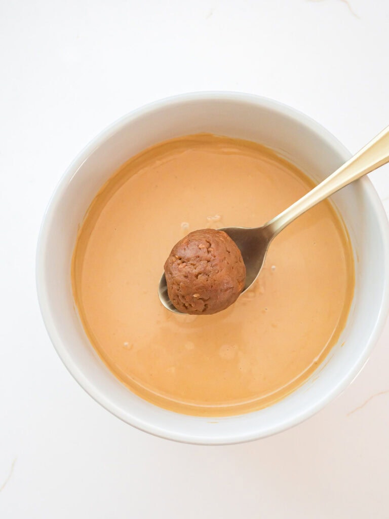 Biscoff truffle ball held on a spoon above a bowl of melted white chocolate and cookie butter mixture for dipping.