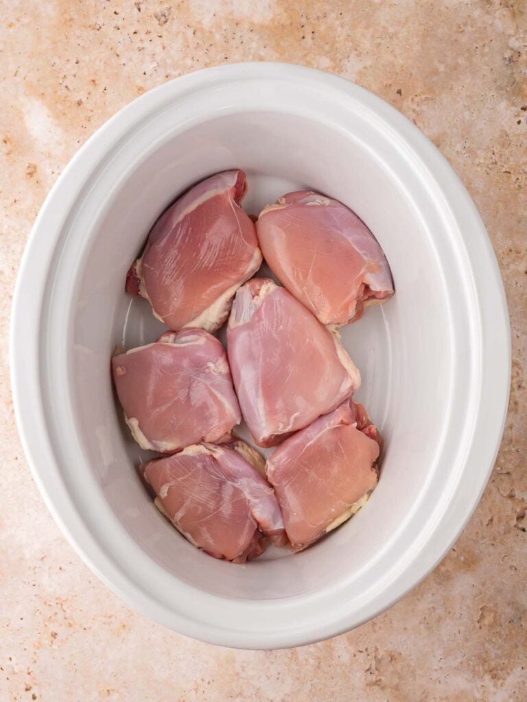 Raw boneless, skinless chicken thighs arranged in the bottom of a white slow cooker before sauce is added.
