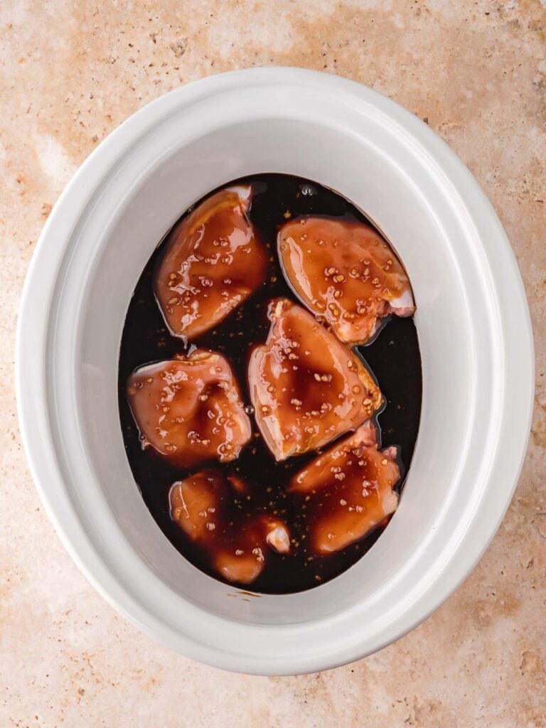 Raw chicken thighs coated in honey garlic sauce before cooking inside a white crock pot.