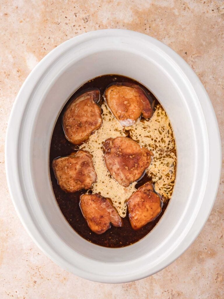 Partially cooked chicken thighs in the slow cooker as the sauce begins to bubble and thicken.