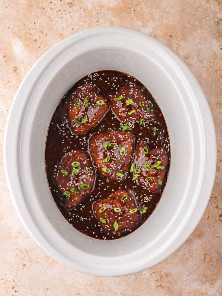 Slow cooker filled with honey garlic chicken thighs topped with sesame seeds and green onions in a thick, glossy sauce.