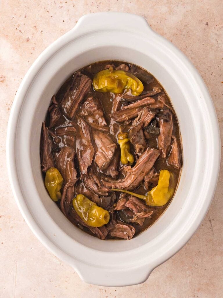 Shredded Slow Cooker Mississippi Pot Roast in white crockpot surrounded by pepperoncini peppers in flavorful gravy.