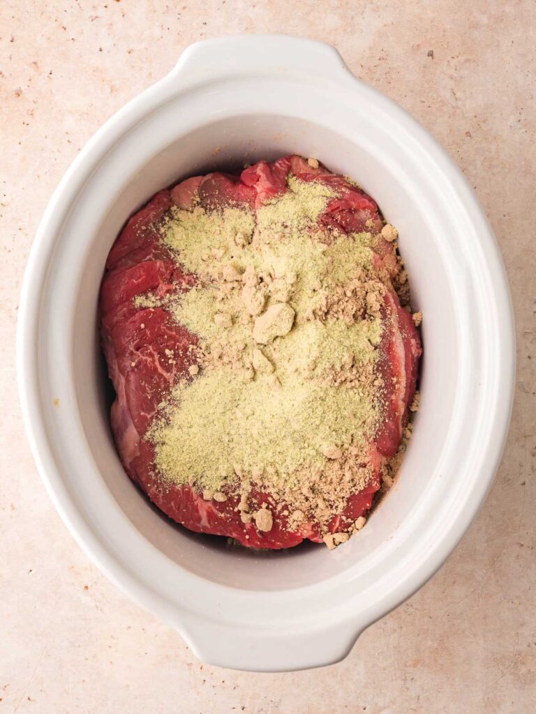 Raw chuck roast in a white slow cooker with dry ranch and au jus seasoning sprinkled on top for Slow Cooker Mississippi Pot Roast.