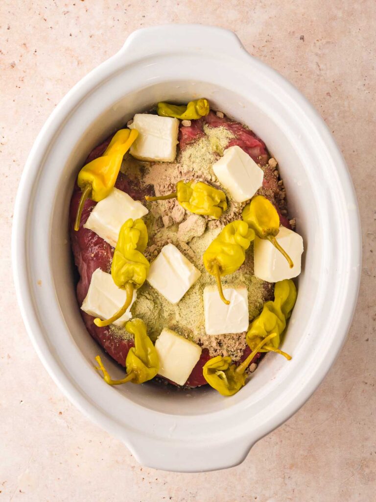 Chuck roast in slow cooker topped with butter cubes, ranch mix, au jus mix, and whole pepperoncini peppers before cooking Mississippi Pot Roast.