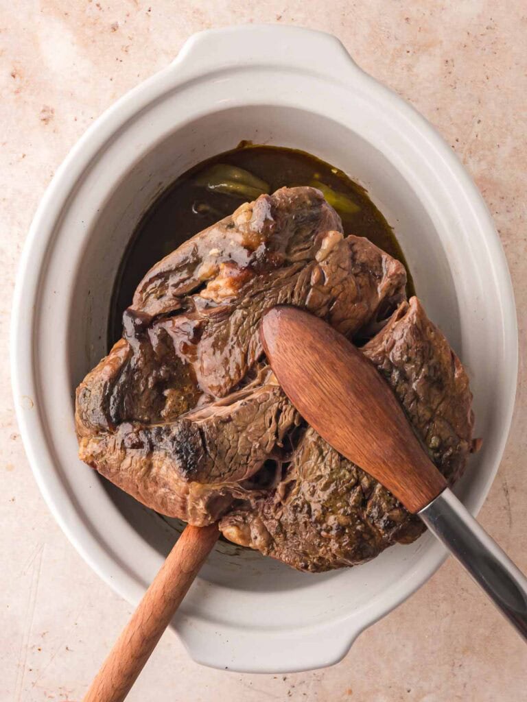 Slow-cooked chuck roast resting in slow cooker with wooden utensils lifting tender pieces of Mississippi Pot Roast.