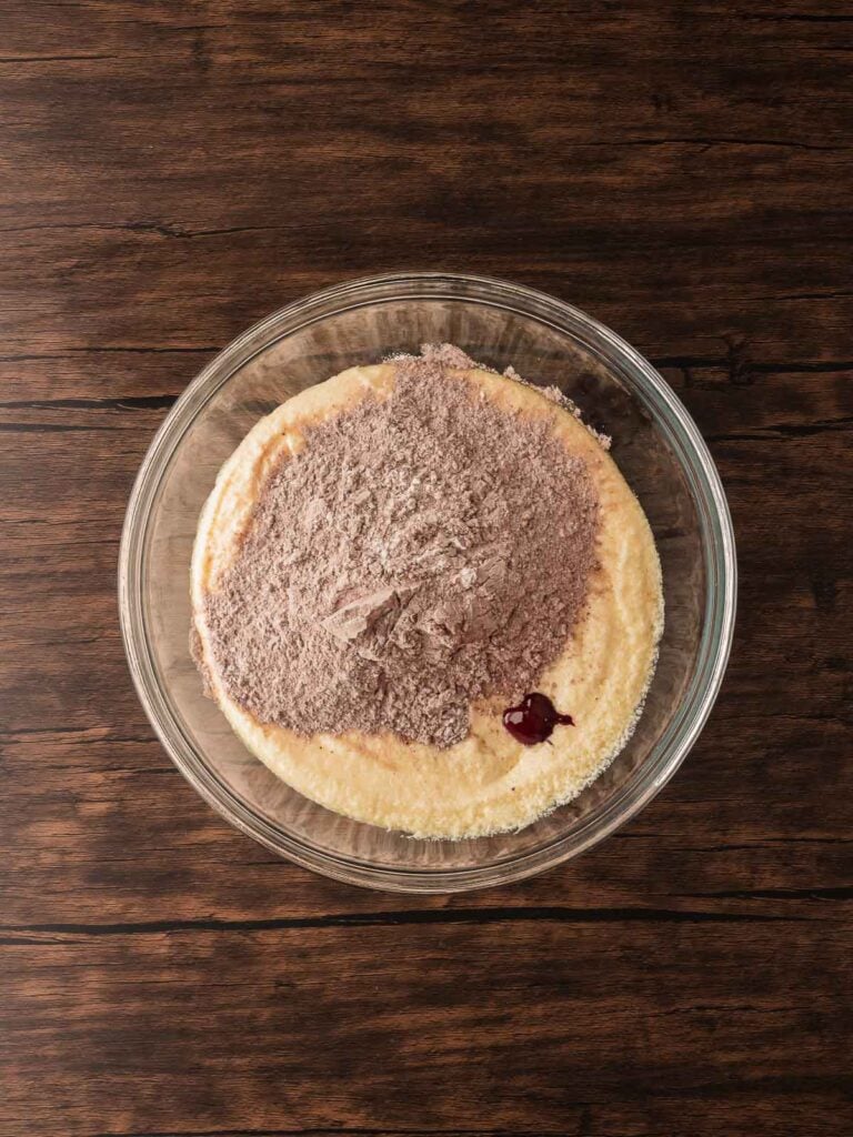 Butter, sugar, eggs, and vanilla creamed together in a glass bowl with cocoa powder and red food coloring added on top for red velvet crinkle cookie dough.