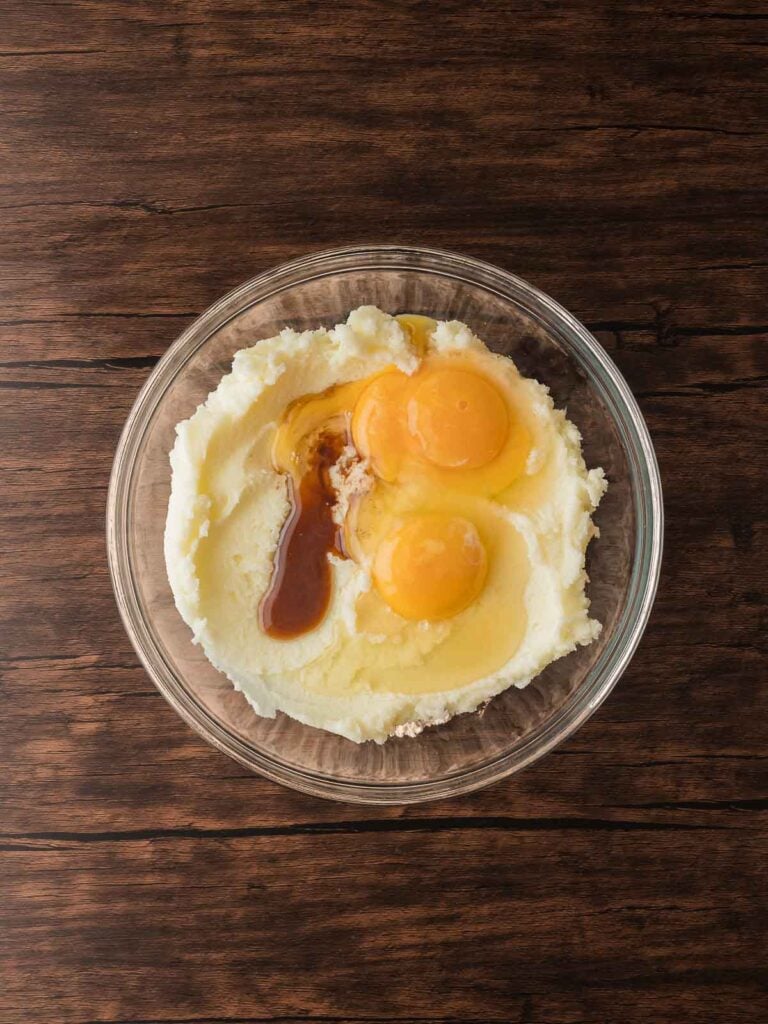 Creamed butter and sugar mixture in a glass bowl with two cracked eggs and vanilla extract before blending for red velvet crinkle cookies.