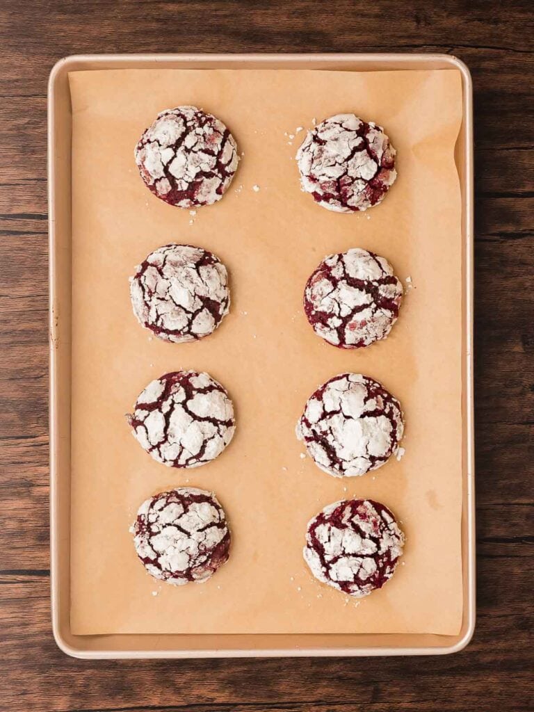 Baked red velvet crinkle cookies on a parchment-lined baking sheet showing crackled powdered sugar tops.