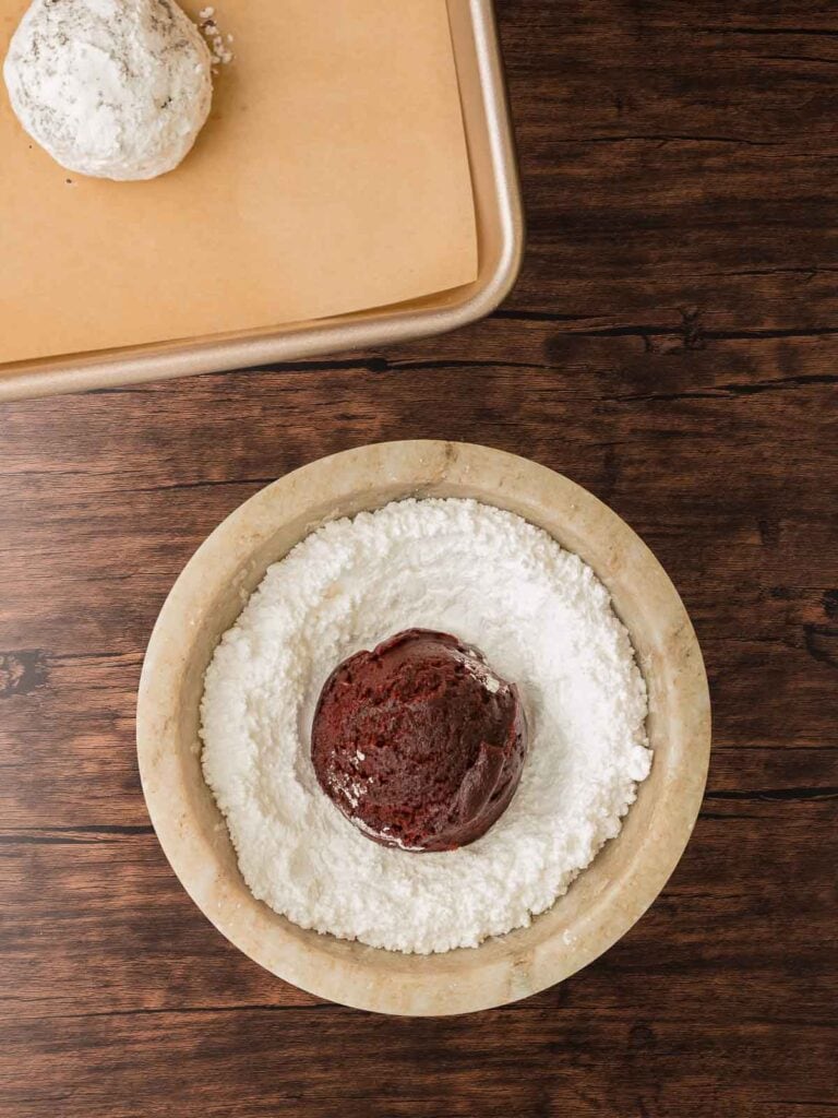 A ball of red velvet cookie dough being rolled in powdered sugar before baking for classic crinkle texture.