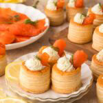 Smoked salmon puff pastry appetizers arranged on a plate and cutting board, with dill cream cheese, lemon slices, and extra smoked salmon displayed around them.
