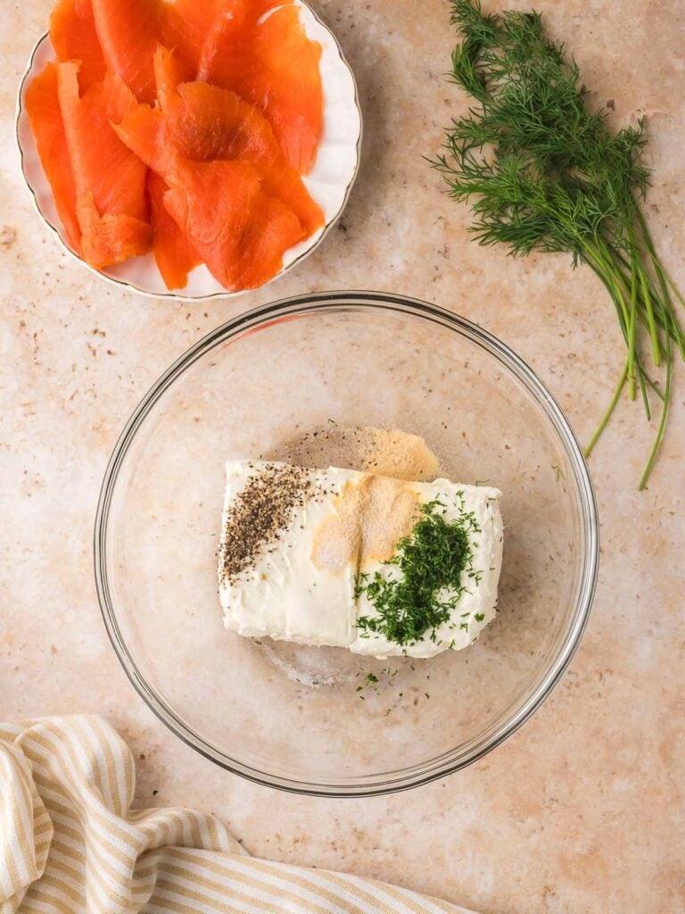 Smoked salmon slices on a plate beside a bowl of cream cheese, dill, garlic powder, and pepper ready to be mixed.