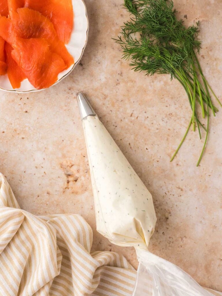 Piping bag filled with dill cream cheese lying beside fresh dill and slices of smoked salmon.