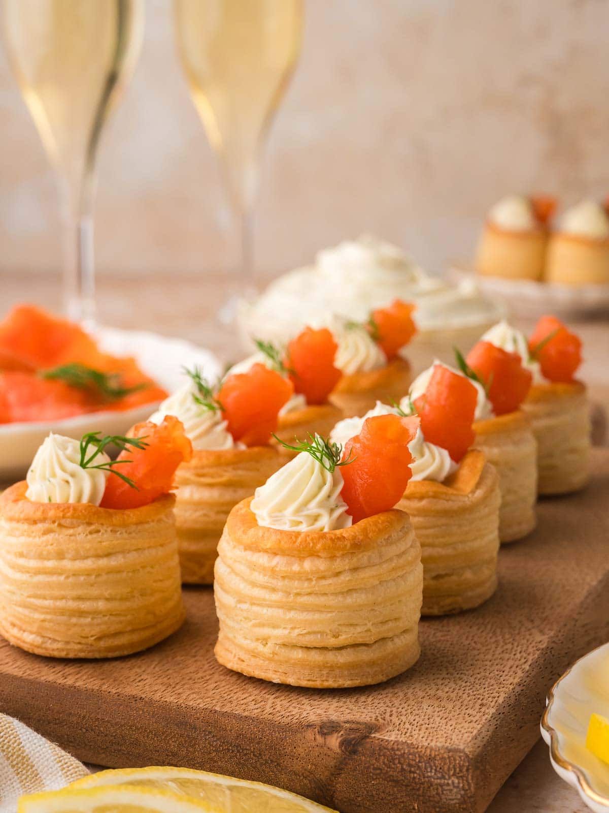 Row of smoked salmon puff pastry appetizers arranged on a wooden board with champagne glasses in the background.